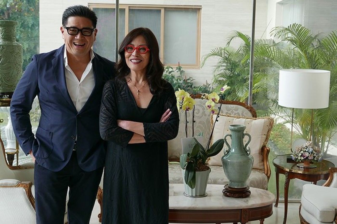 Anton Huang in a navy suit laughs beside Cecile Licad, in black with red-framed glasses, before a sunlit window with orchids