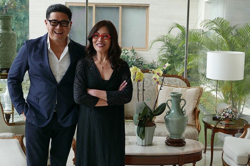 Anton Huang in a navy suit laughs beside Cecile Licad, in black with red-framed glasses, before a sunlit window with orchids