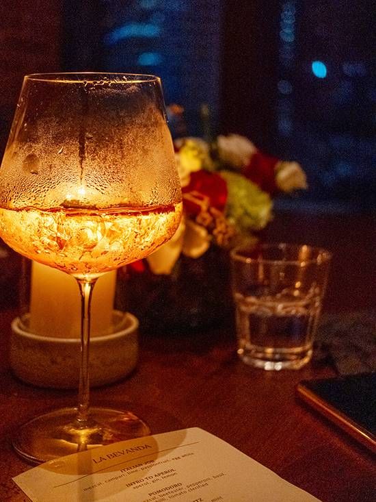 Amber cocktail in a stemmed glass glowing above candlelight beside flowers and Osteria Antica’s La Bevanda drinks menu