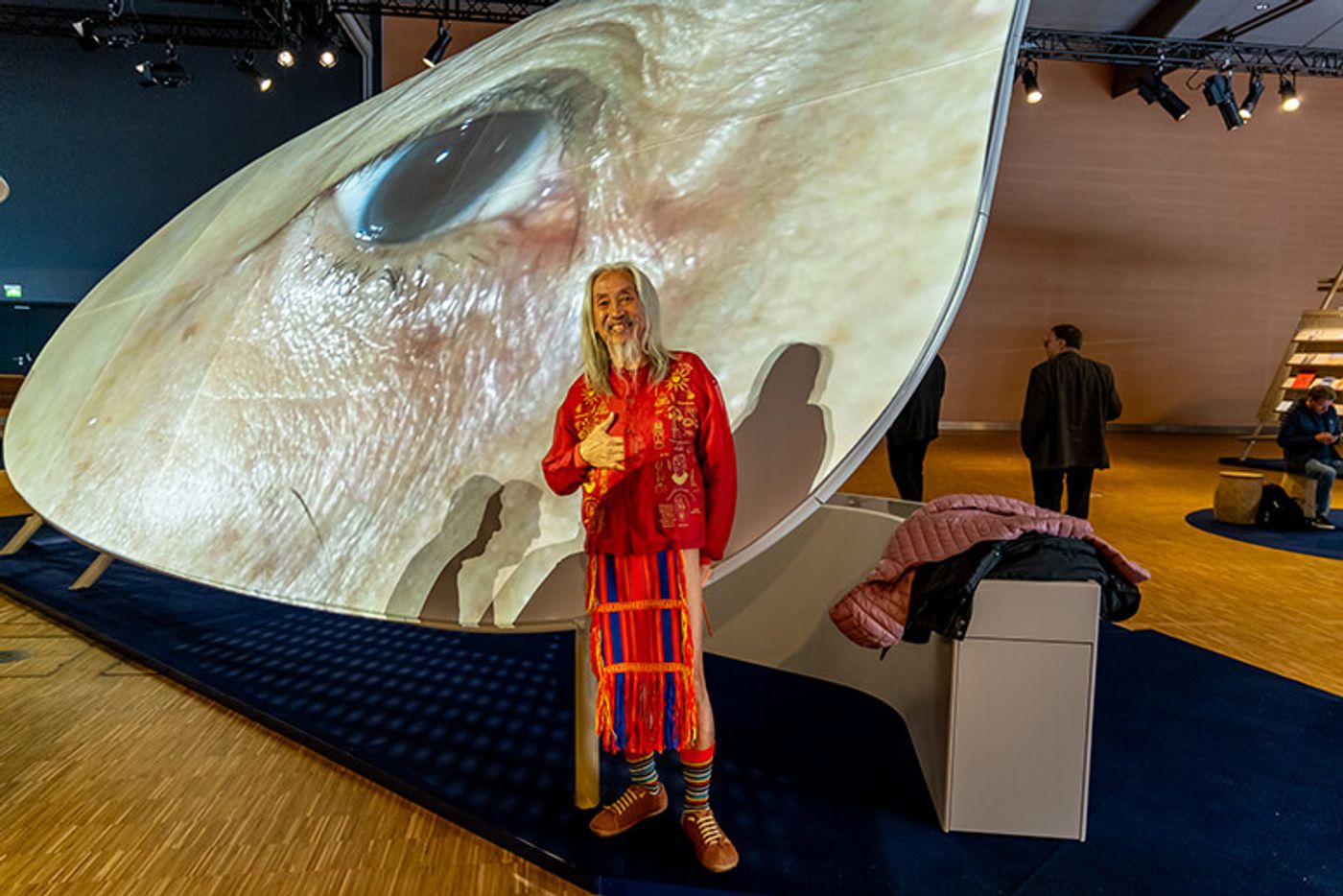 National Artist Kidlat Tahimik in red traditional dress giving a thumbs up beneath a giant projected eye