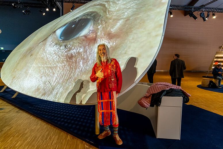 National Artist Kidlat Tahimik in red traditional dress giving a thumbs up beneath a giant projected eye