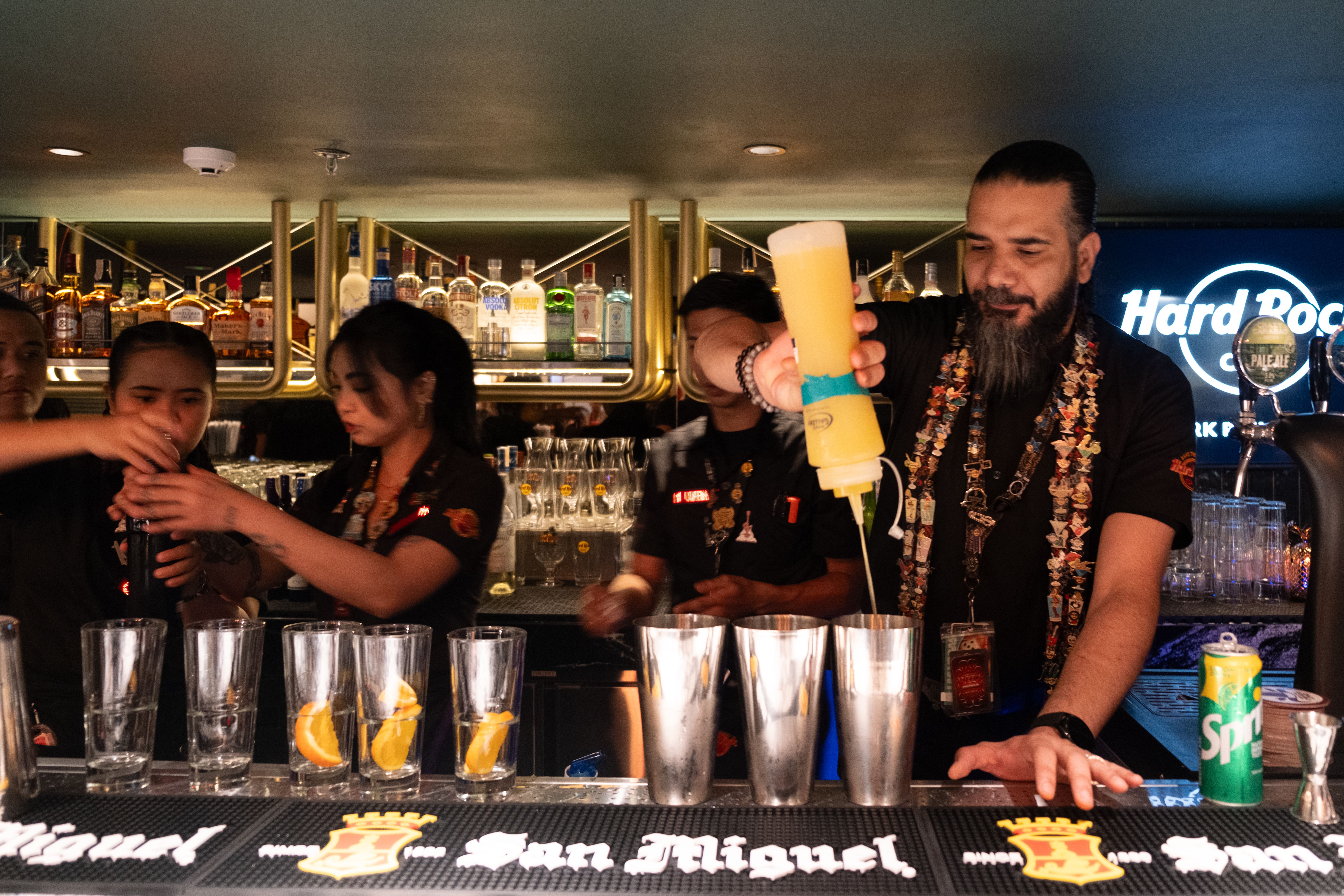 A bearded bartender in a pin-covered lanyard squeezes yellow cocktail mix into shakers at the Hard Rock Cafe Clark bar