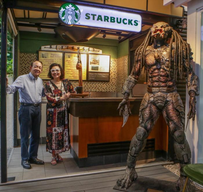 Jun and Menchu Lopez beside a home coffee bar built like a Starbucks counter, with a life-size Predator figure standing guard at the right