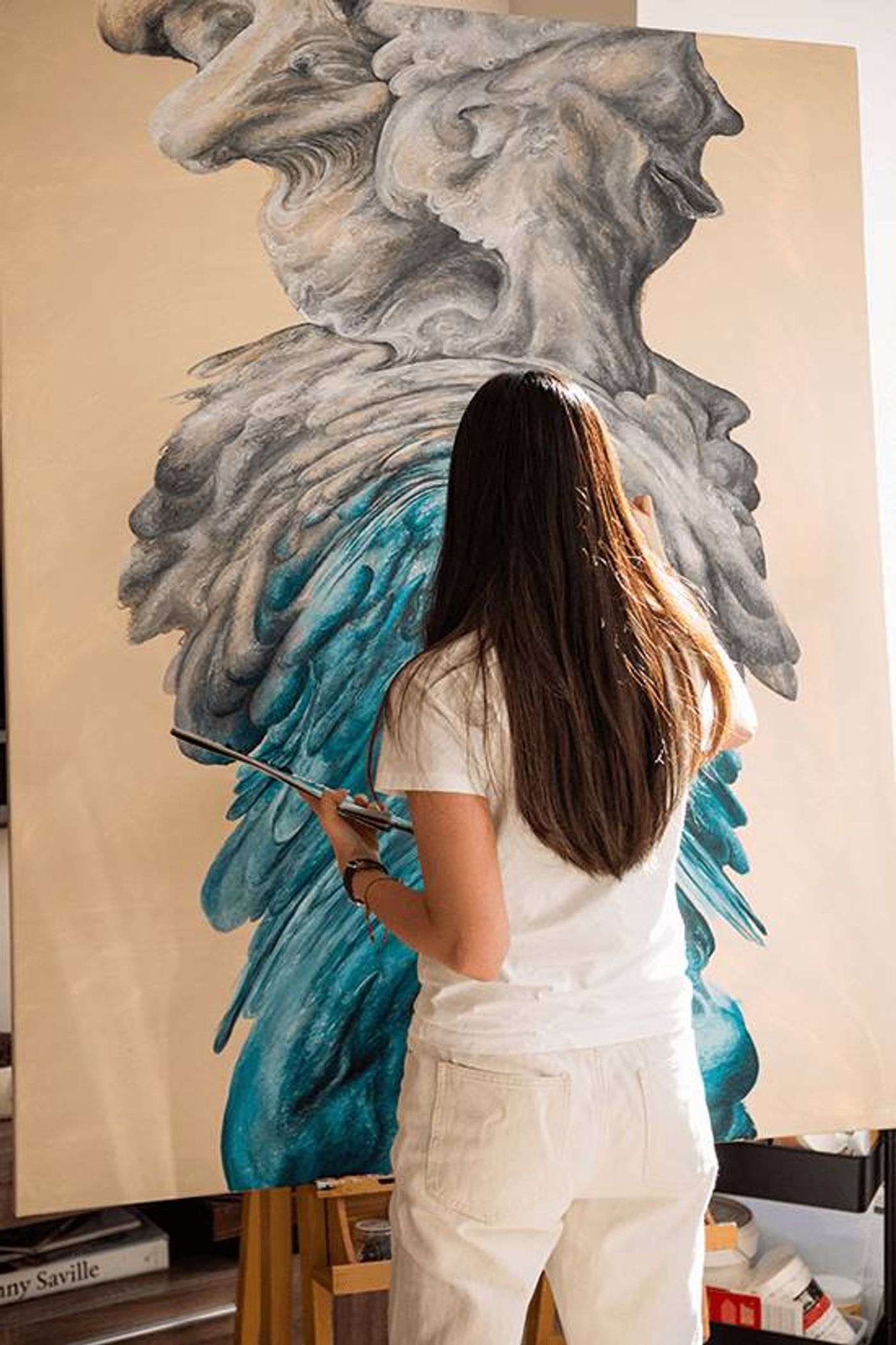 Maria Angelica Tan paints a tall canvas of grey and turquoise cloudlike forms in her studio
