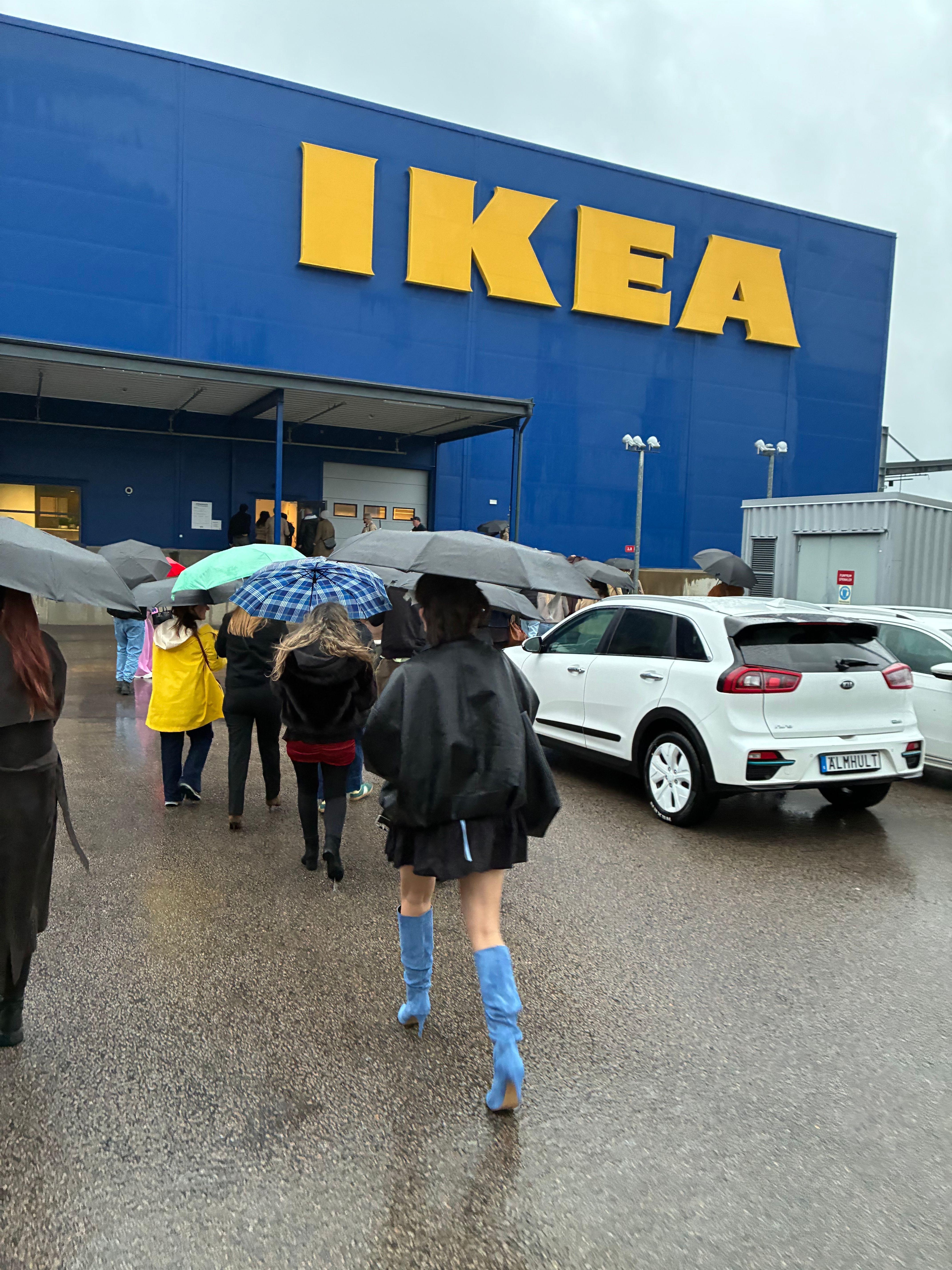 Guests with umbrellas cross a rainy car park toward the blue and yellow IKEA store facade in Älmhult