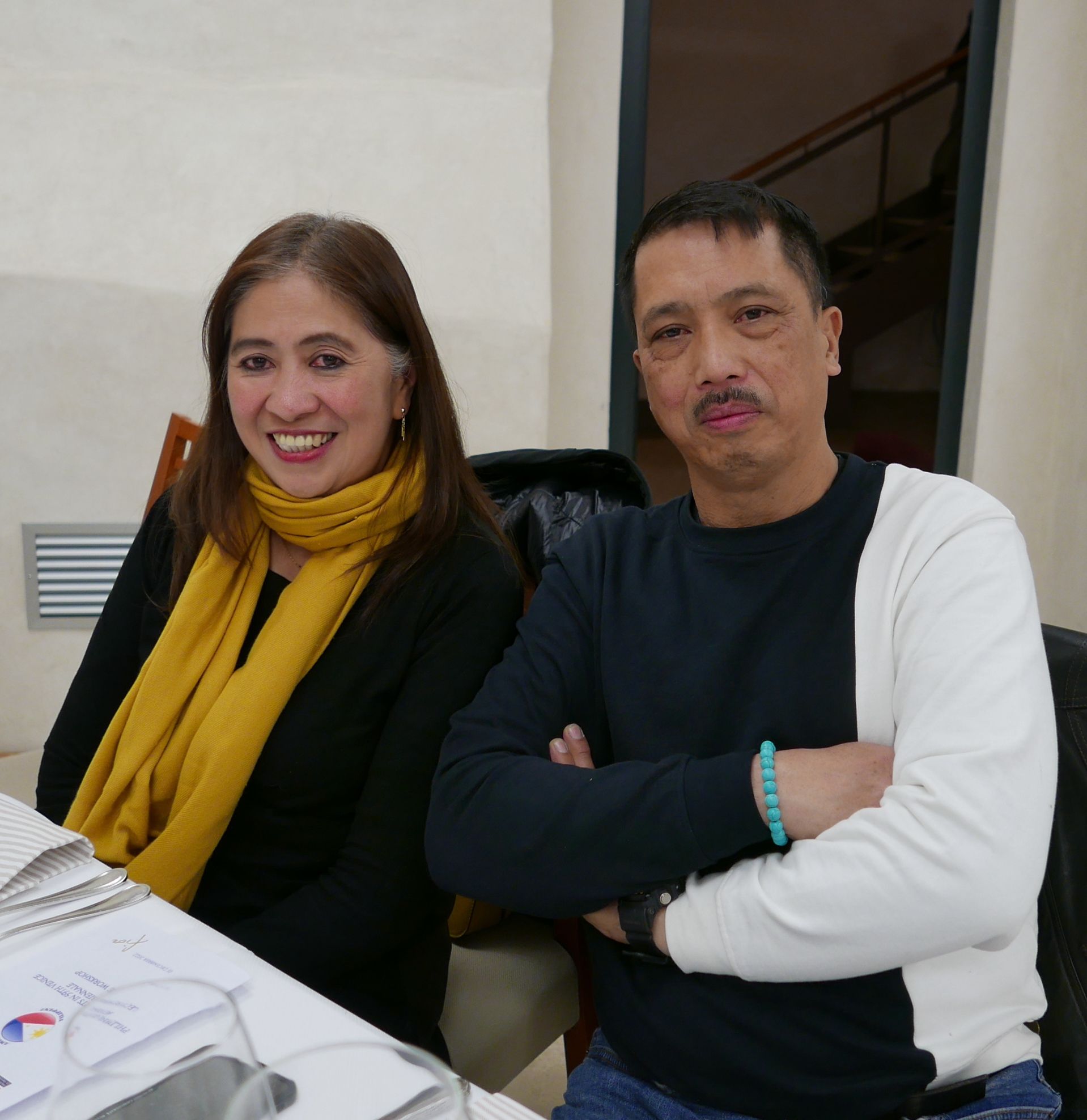 Linda and Ramon Lagpao sit together at a table, she smiling in a mustard scarf, he in a black-and-white sweater with arms crossed