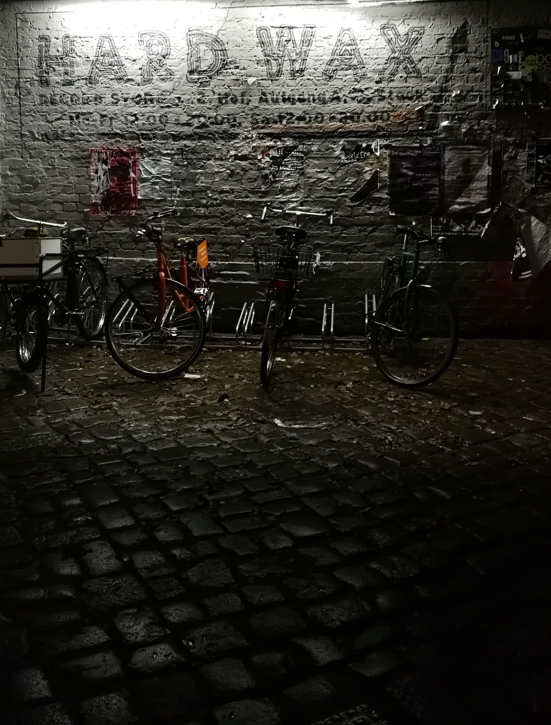 Bicycles parked on wet cobblestones at night below the painted Hard Wax sign on a brick wall