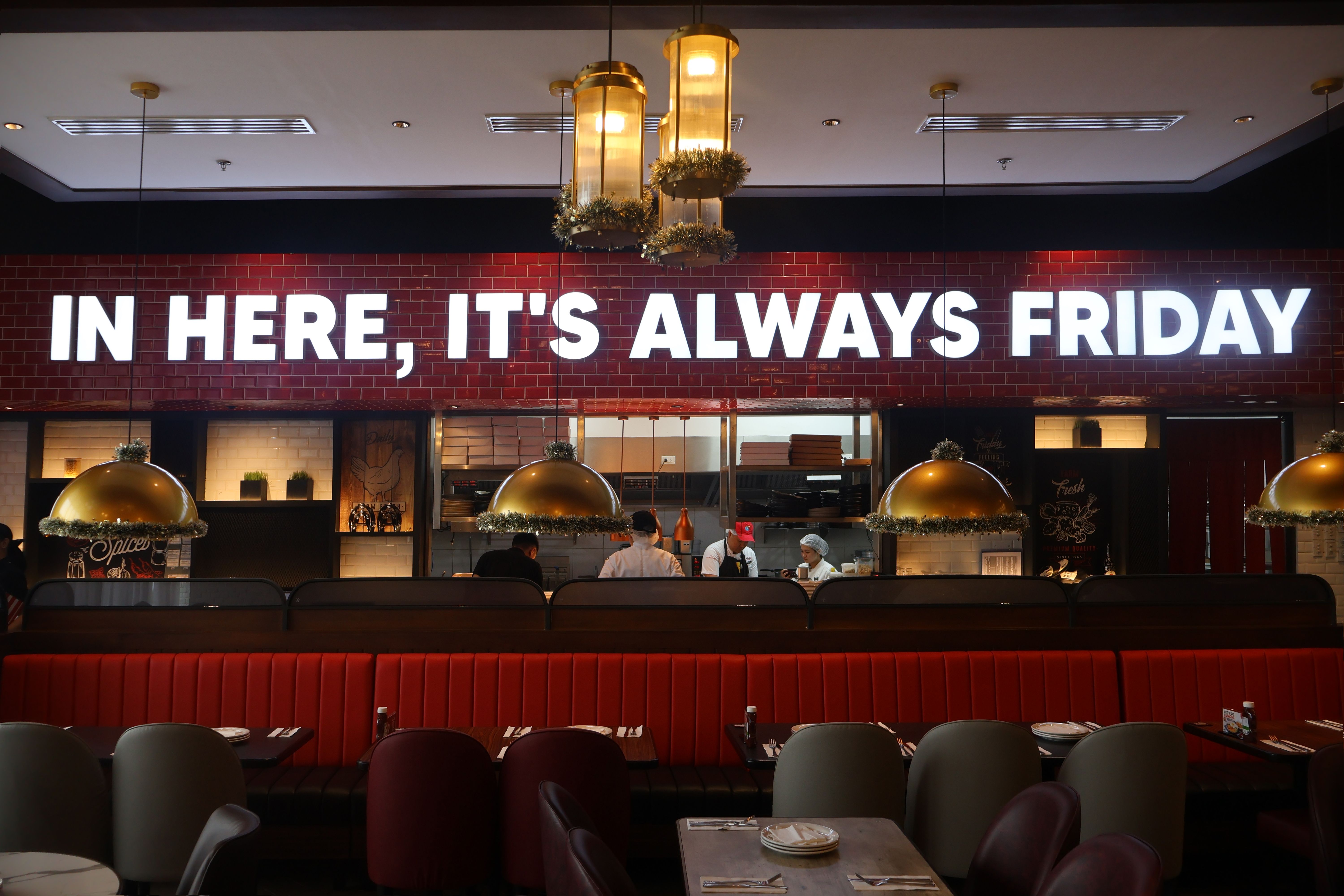 Red banquettes and brass dome lamps beneath a glowing sign reading In here, it’s always Friday