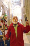 A white-bearded pilgrim in a red jacket raising a book and a ribboned staff inside the Santiago Cathedral