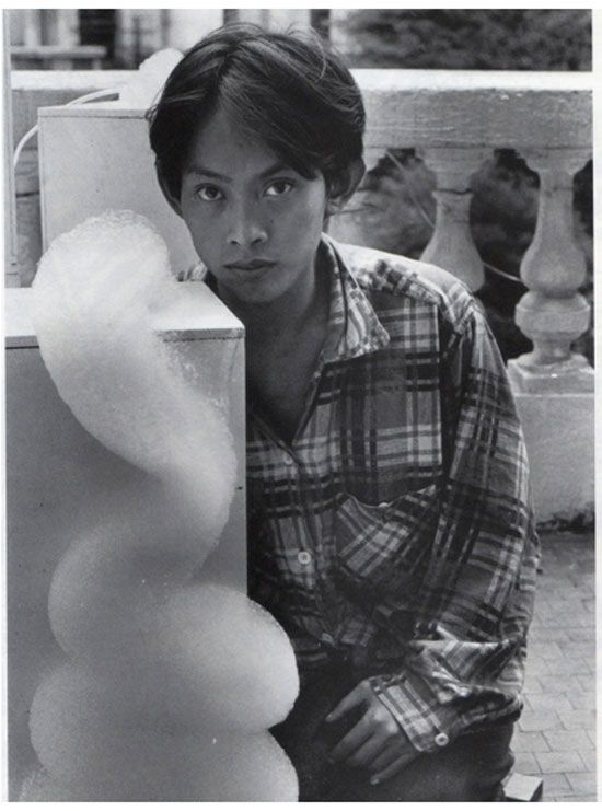 Black and white portrait of a young David Medalla in a plaid shirt beside a column of foam from a bubble machine