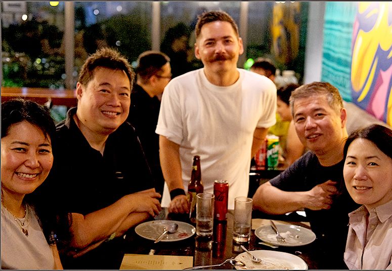 Chef Josh Boutwood stands behind a table of grinning friends at a rooftop restaurant