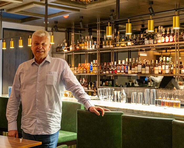 John W. Hardyment leans on a green banquette before a backlit bar stacked with whisky bottles