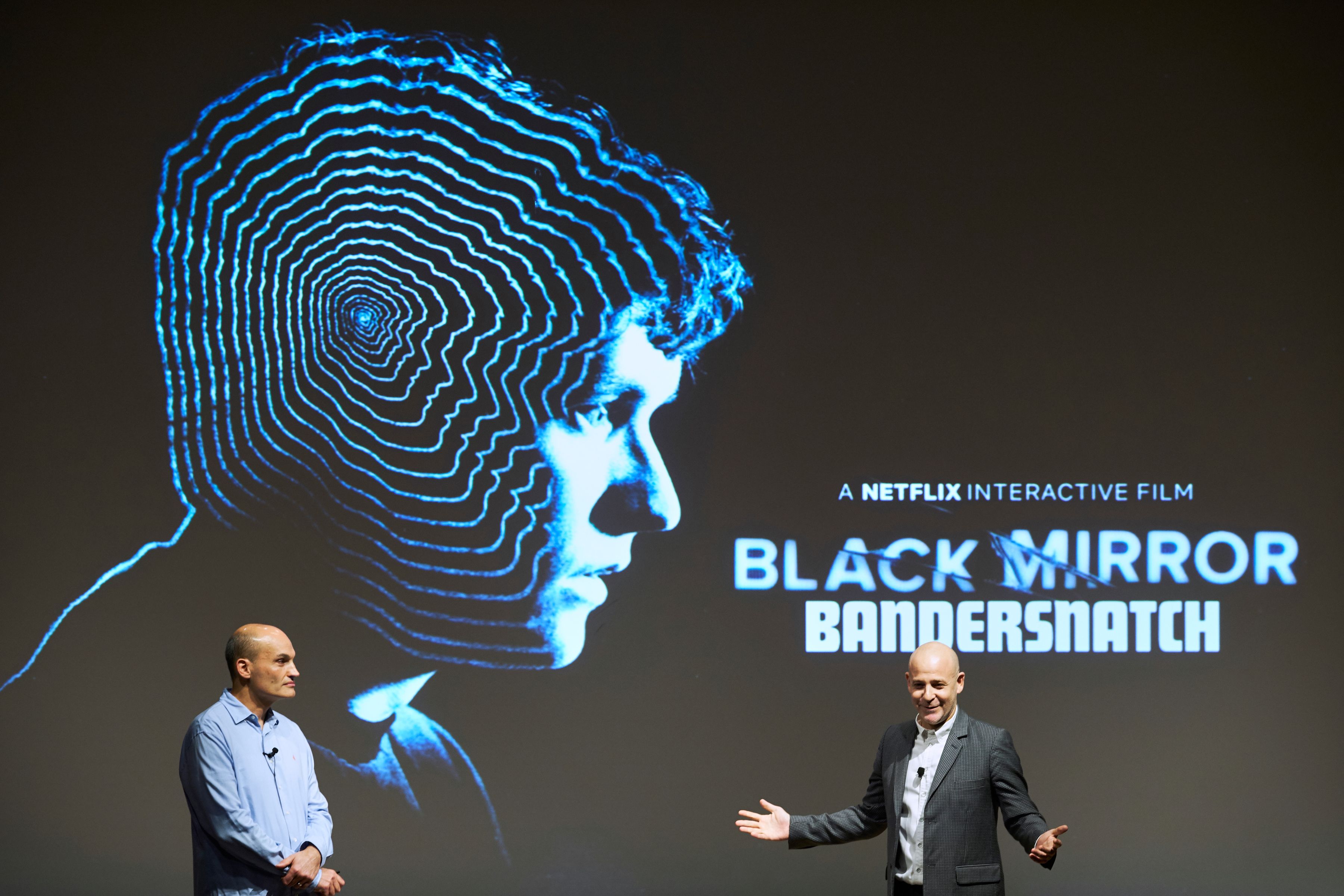 Two presenters onstage beside the glowing spiral-head key art for Black Mirror: Bandersnatch