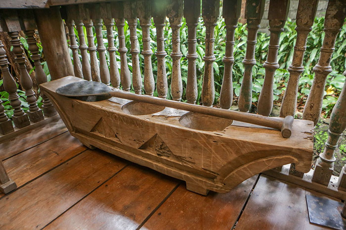 A lusong rice mortar carved from a single log, its wooden pestle laid across it, on the plank floor of the bahay kubo behind turned balusters