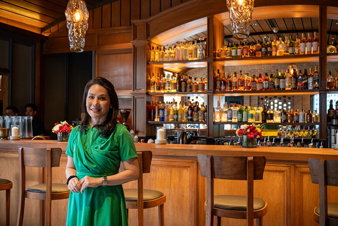 Osteria Antica’s Ana Lorenzana de Ocampo in a green dress standing before the restaurant’s stocked wooden bar