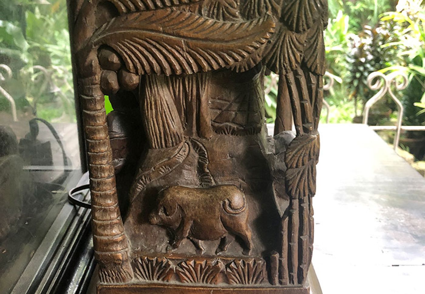 A carved wooden relief of a carabao standing under coconut palms, the panel set against a garden window