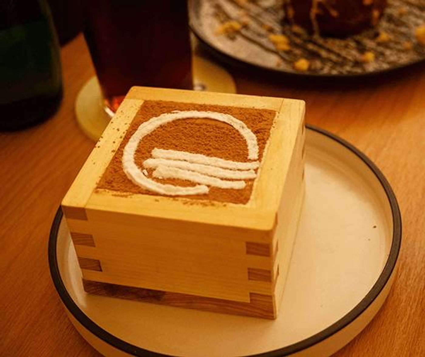 Classic tiramisu dusted with cocoa in a wooden masu box, a plated lava cake behind it