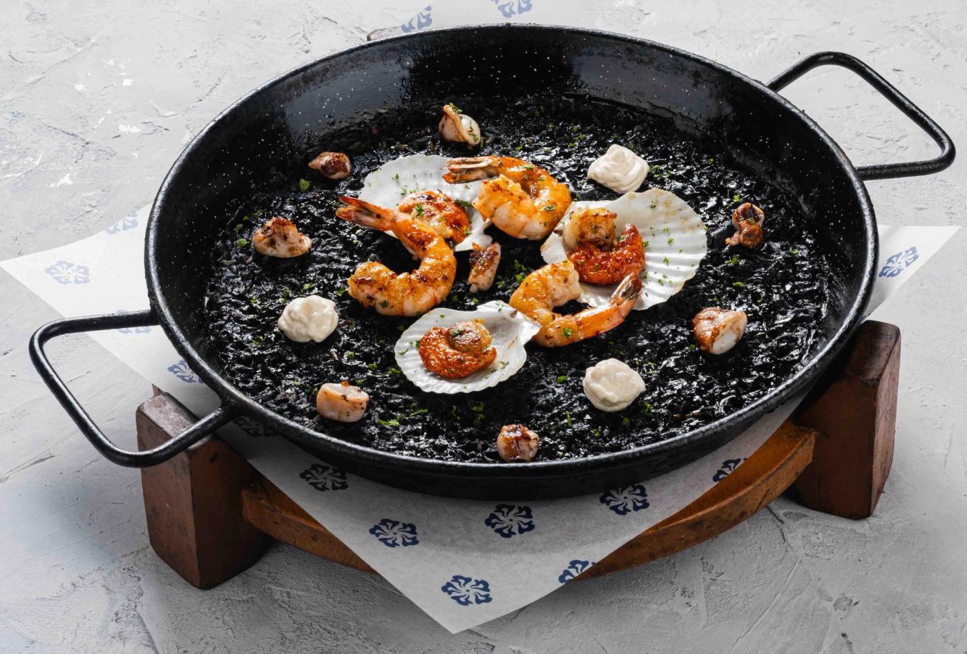Paella Negra in a black steel pan, squid-ink rice topped with grilled shrimp, scallops in shells and dollops of aioli
