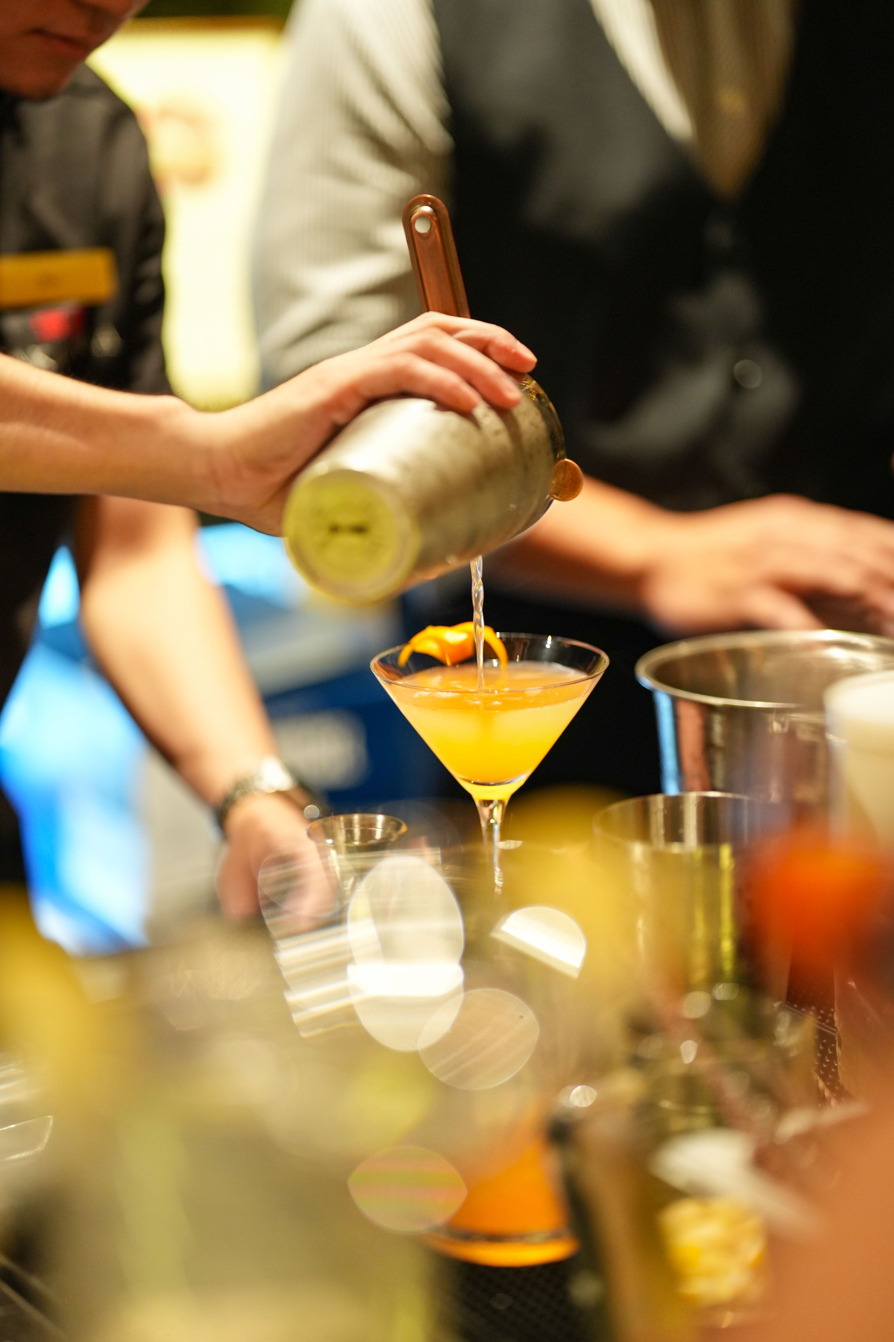 A bartender strains a golden orange cocktail from a shaker into a martini glass garnished with an orange twist