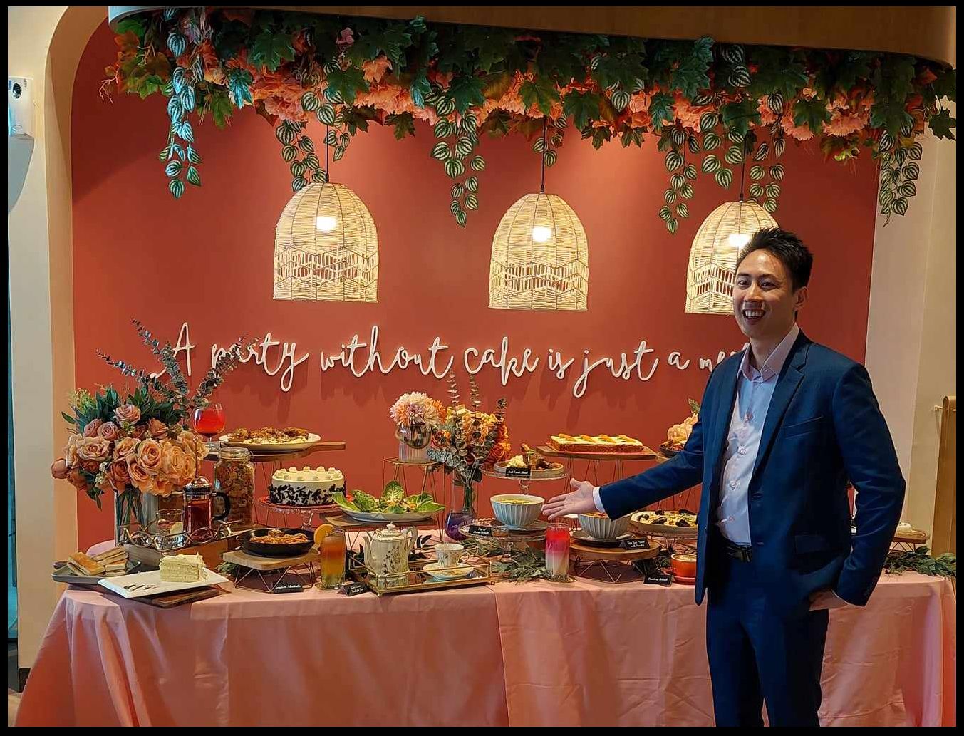 Patrick Sim in a blue suit gestures toward a lavish dessert table of cakes, teapots and flowers, beneath a terracotta wall with birdcage lamps and neon script reading “A party without cake is just a meeting.”