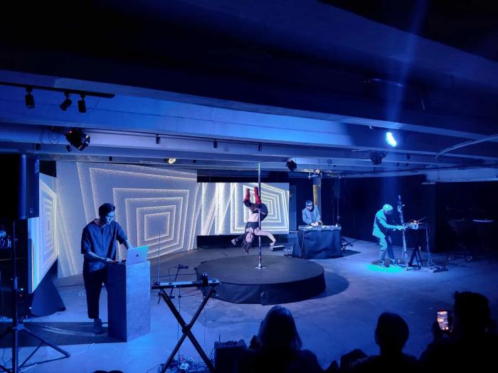 Wide shot of Somatosonic performing Totality 1.0 under blue stage lights: Christina Dy hanging upside-down on a pole, Tad Ermitaño at the electronics table, and Marco Ortiga bowing the Tungkod