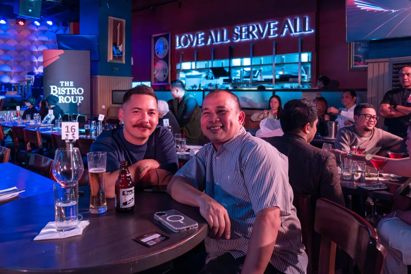 Chef Josh Boutwood and William Abuel share a table beneath the neon Love All Serve All sign