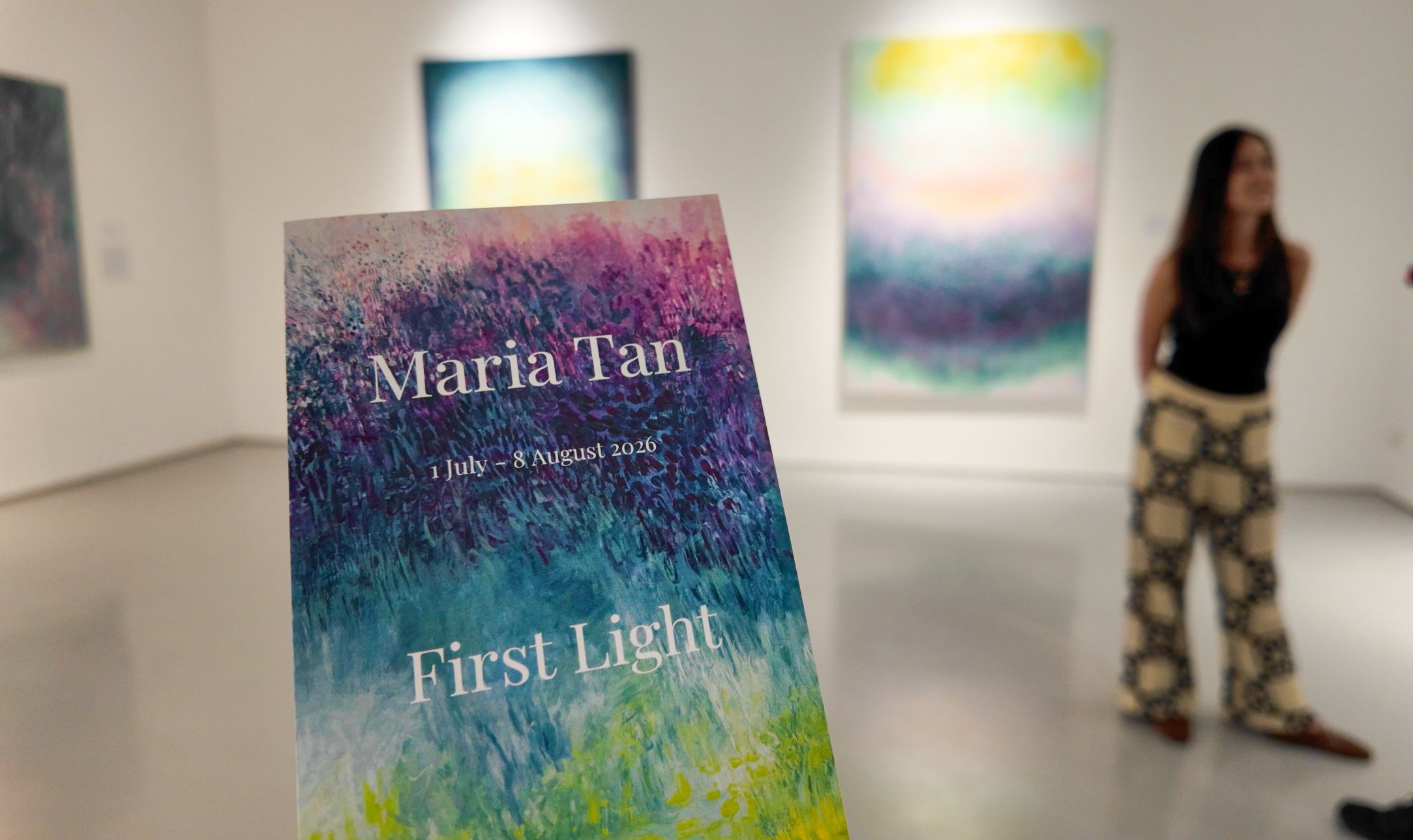 A First Light show card in the foreground with Maria Tan standing among her paintings at The Columns Gallery