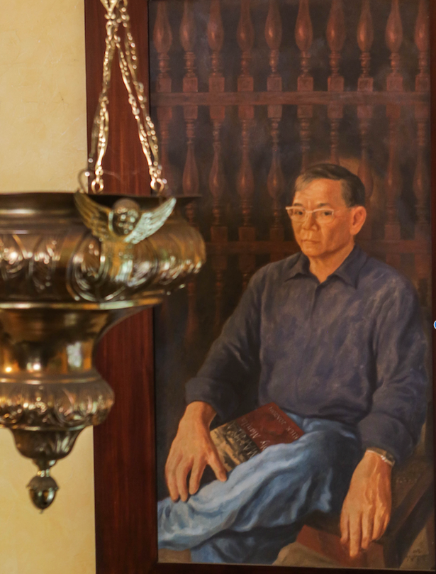 An oil portrait of Jaime Laya seated in a blue shirt with a book on his lap, hung in a dark wooden frame beside an ornate brass lamp