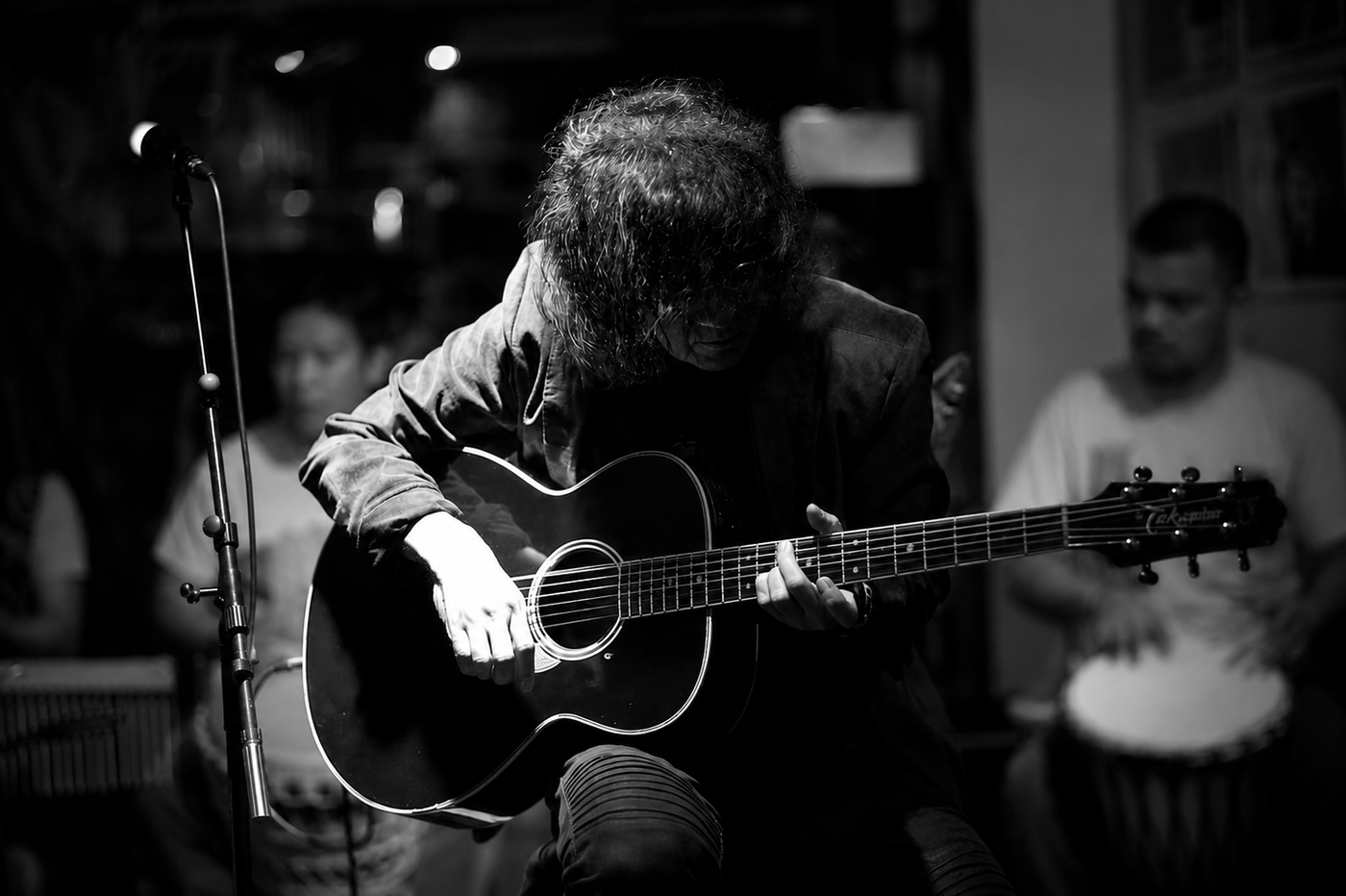 In a black-and-white photo, guitarist Noli Aurillo bends low over his acoustic guitar, hair veiling his face, playing in a dim bar beside a microphone stand