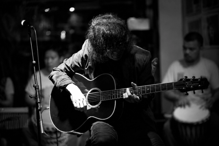 In a black-and-white photo, guitarist Noli Aurillo bends low over his acoustic guitar, hair veiling his face, playing in a dim bar beside a microphone stand