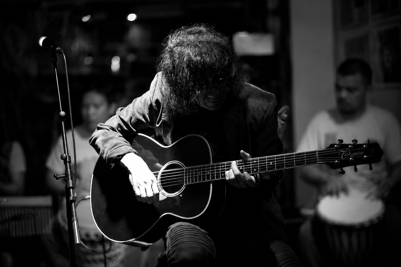 In a black-and-white photo, guitarist Noli Aurillo bends low over his acoustic guitar, hair veiling his face, playing in a dim bar beside a microphone stand