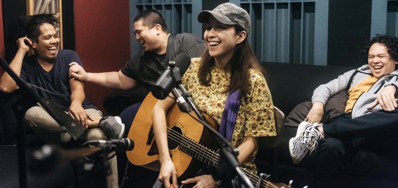 Barbie Almalbis laughing with an acoustic guitar on a studio sofa, her three bandmates around her mid-laugh