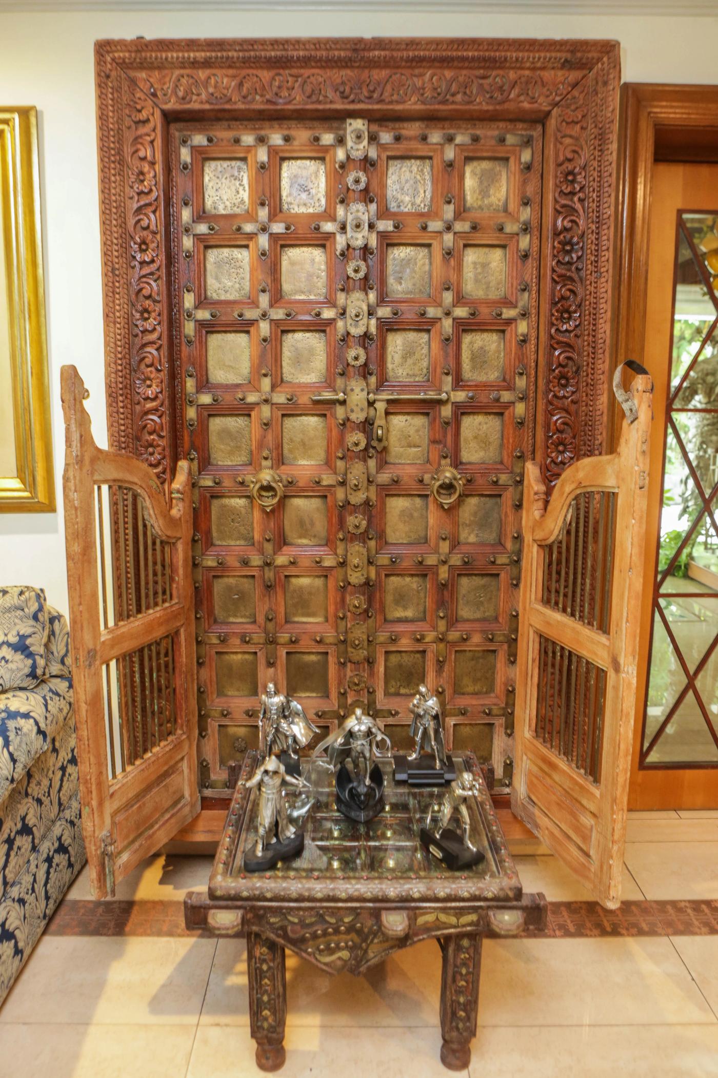 A tall carved Indian door with brass panels and studs, flanked by open shutters, with silver superhero figurines on a low glass table