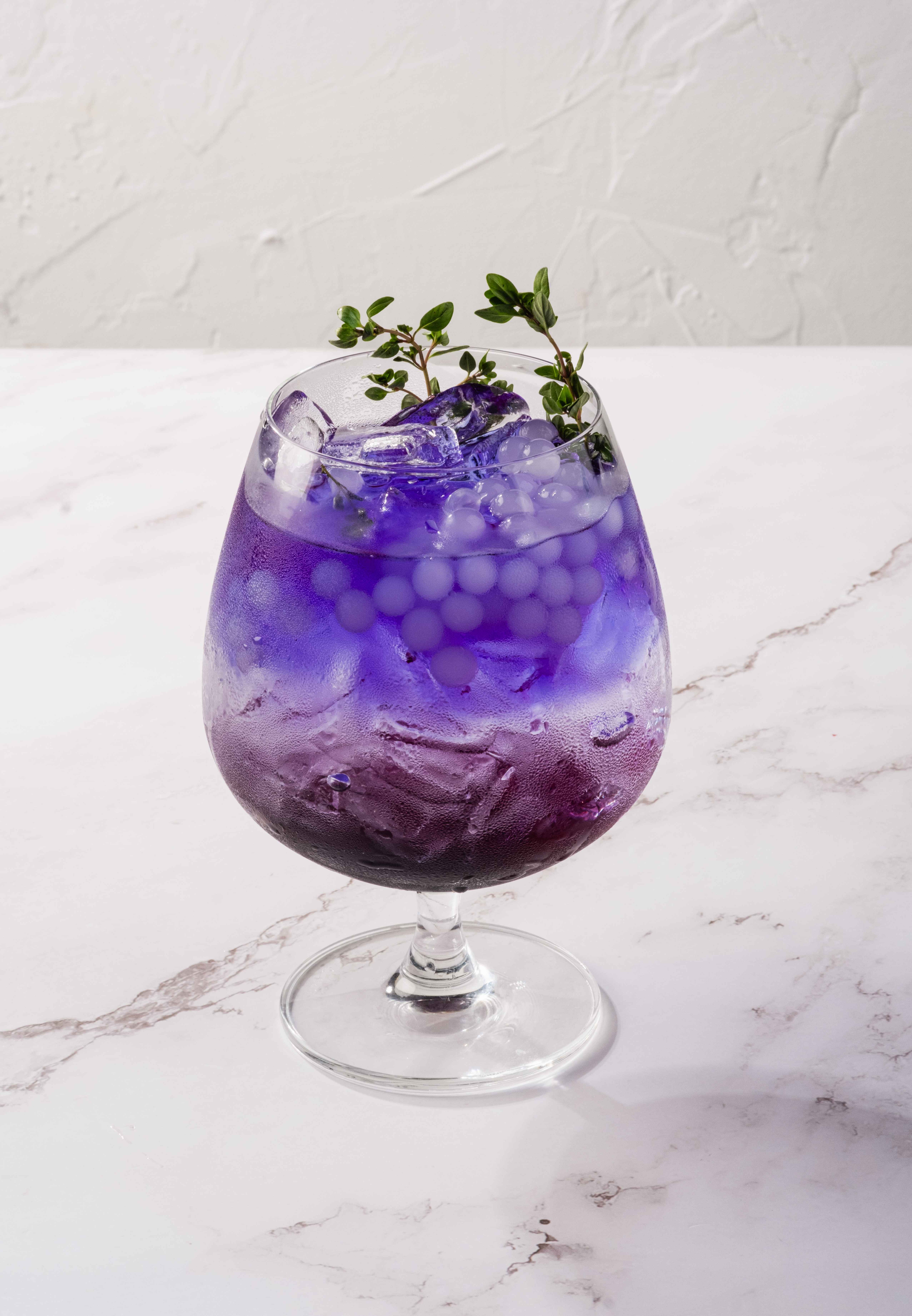 A stemmed glass of iced purple butterfly-pea ‘Citrus Sea of Blue’ tea with jelly pearls, garnished with fresh thyme