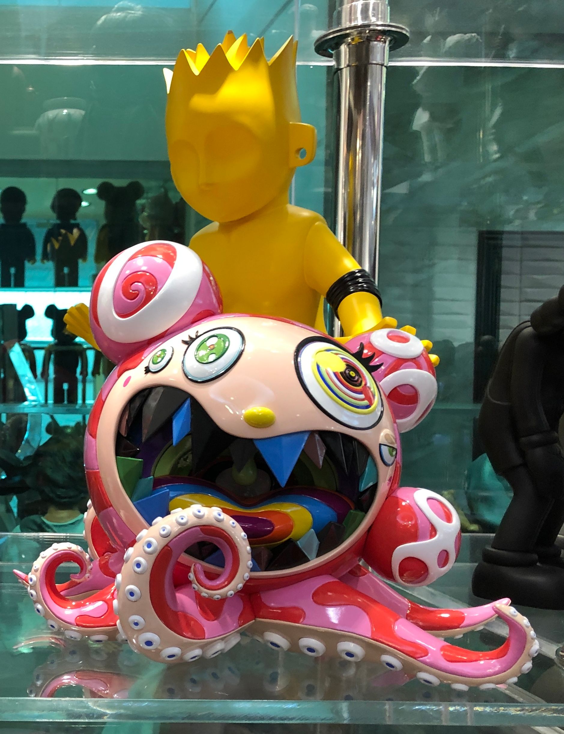 Takashi Murakami’s grinning pink octopus flower-ball in a vitrine with a yellow crowned Bart figure behind it and Bearbricks reflected in the glass