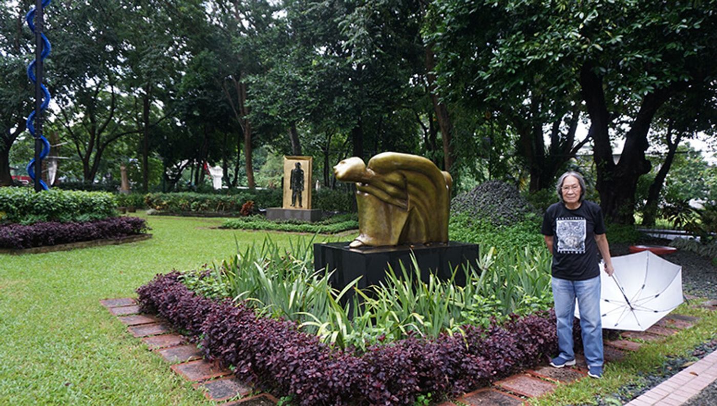 Junyee with an umbrella beside a golden crouching figure at the UP Los Banos Sculpture Garden