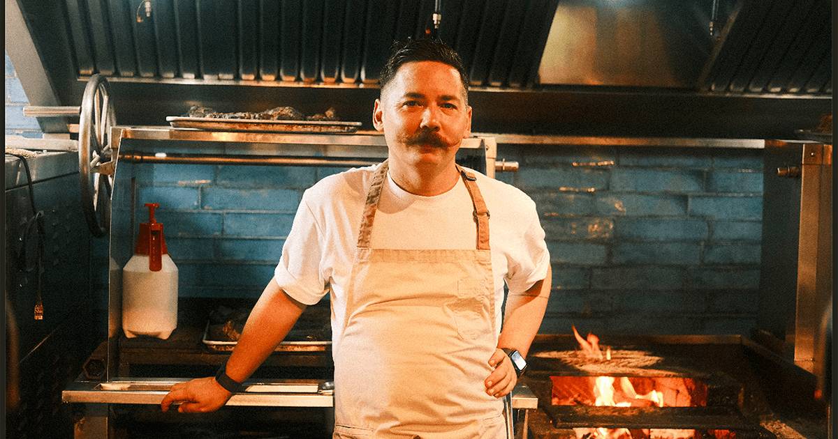 Chef Josh Boutwood, in a white t-shirt and canvas apron with a handlebar mustache, stands before the blue-tiled open-fire grill of his kitchen