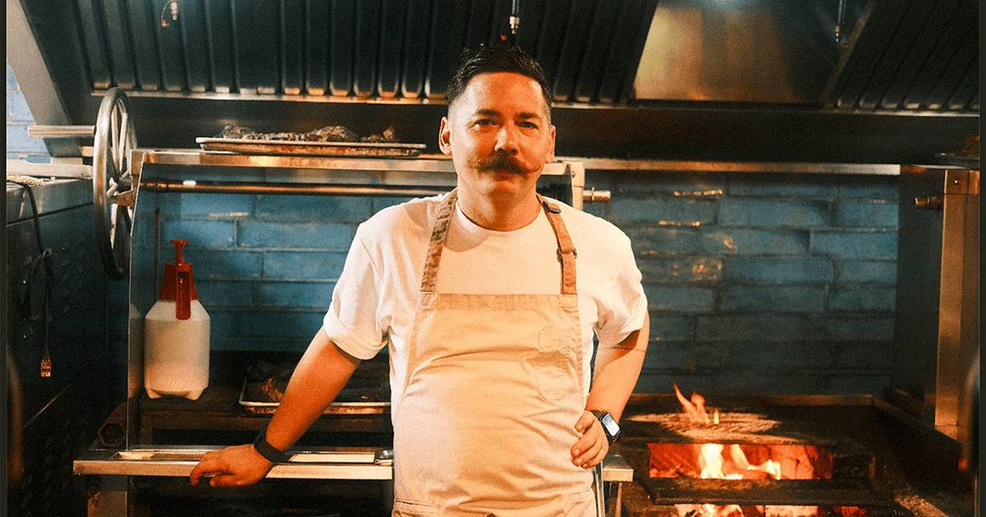 Chef Josh Boutwood, in a white t-shirt and canvas apron with a handlebar mustache, stands before the blue-tiled open-fire grill of his kitchen