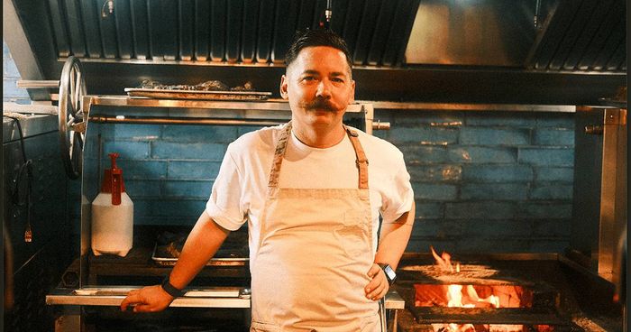 Chef Josh Boutwood, in a white t-shirt and canvas apron with a handlebar mustache, stands before the blue-tiled open-fire grill of his kitchen