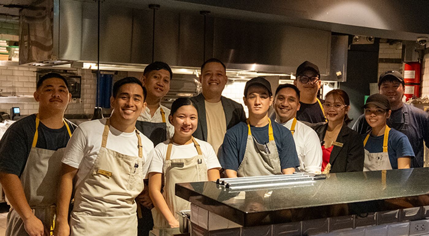 Chef Shanley Ladia and The Test Kitchen crew in aprons posing behind the stainless steel kitchen counter