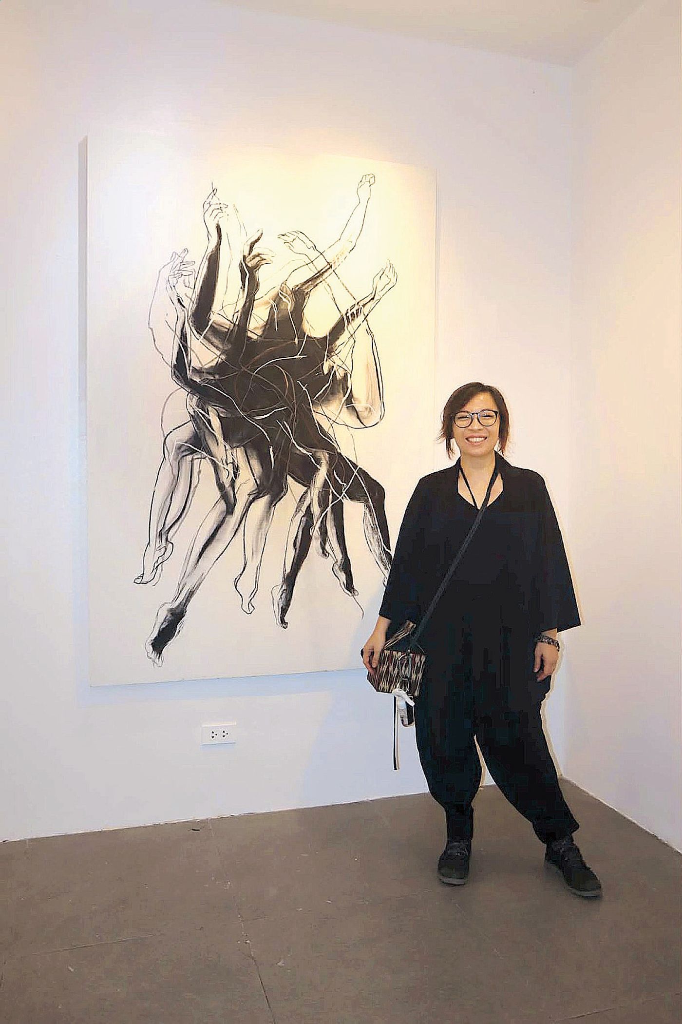 Christina Dy in black beside a large charcoal chimera drawing on a white wall at The Crucible Gallery