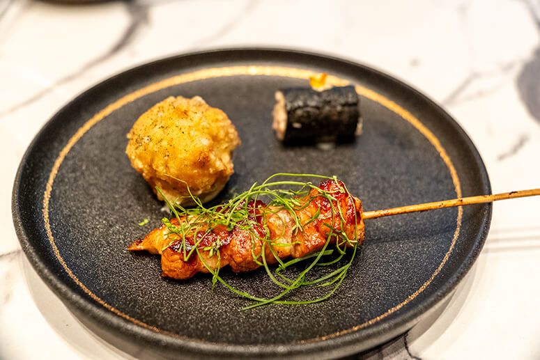 A tamarind-glazed skewer, a piece of Thai fried chicken and a nori-wrapped tuna roll on a dark plate