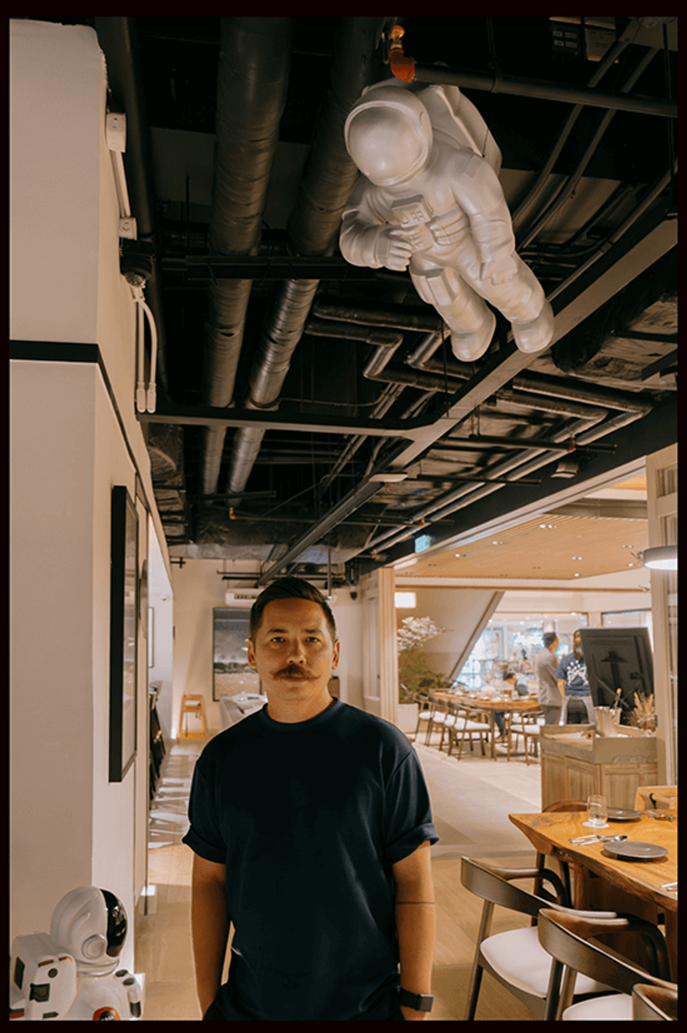 Chef Josh Boutwood stands in his dining room beneath a white astronaut figure suspended from the black ceiling