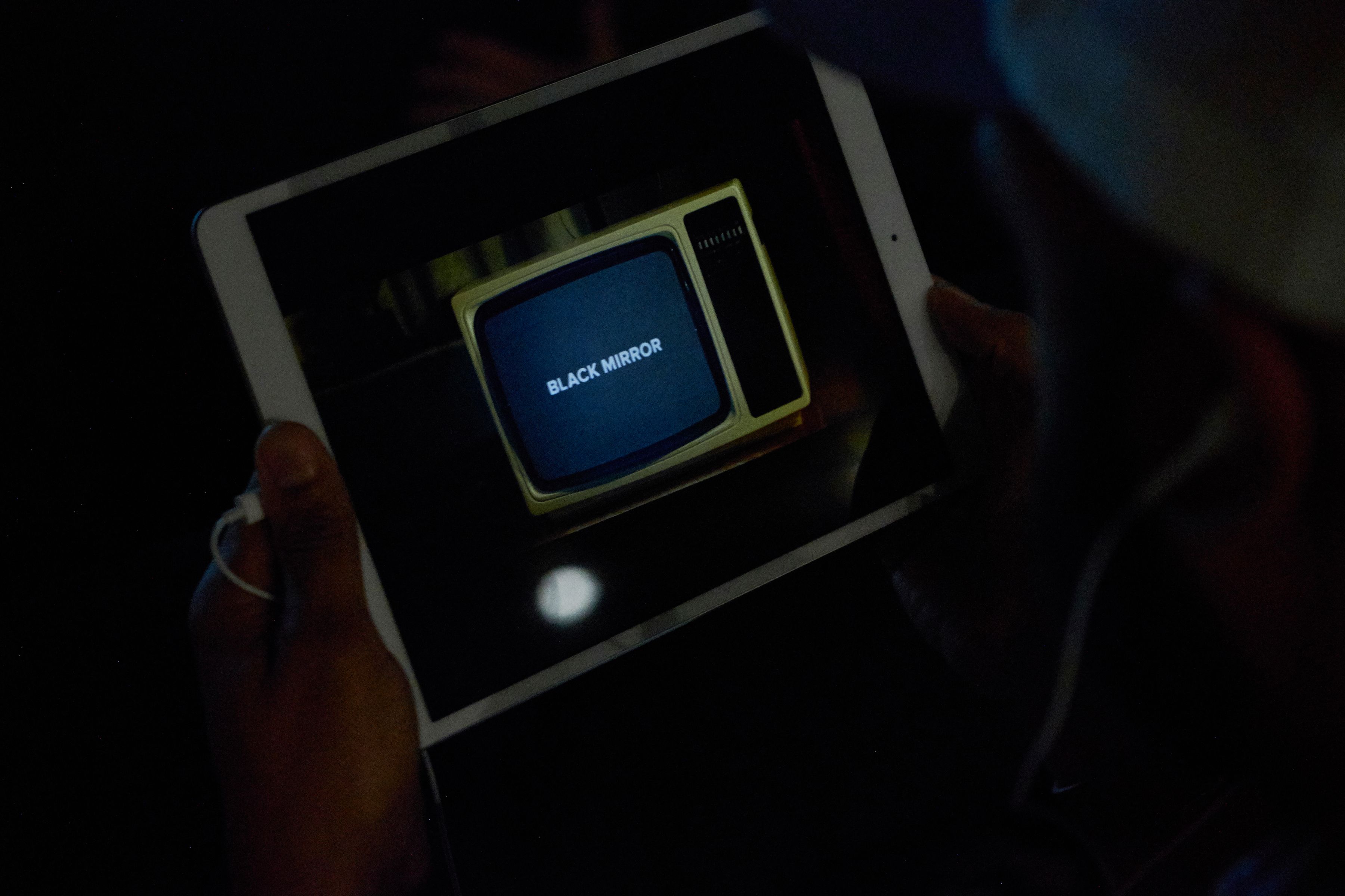 Hands hold a tablet showing a vintage yellow TV set with the Black Mirror logo on its screen
