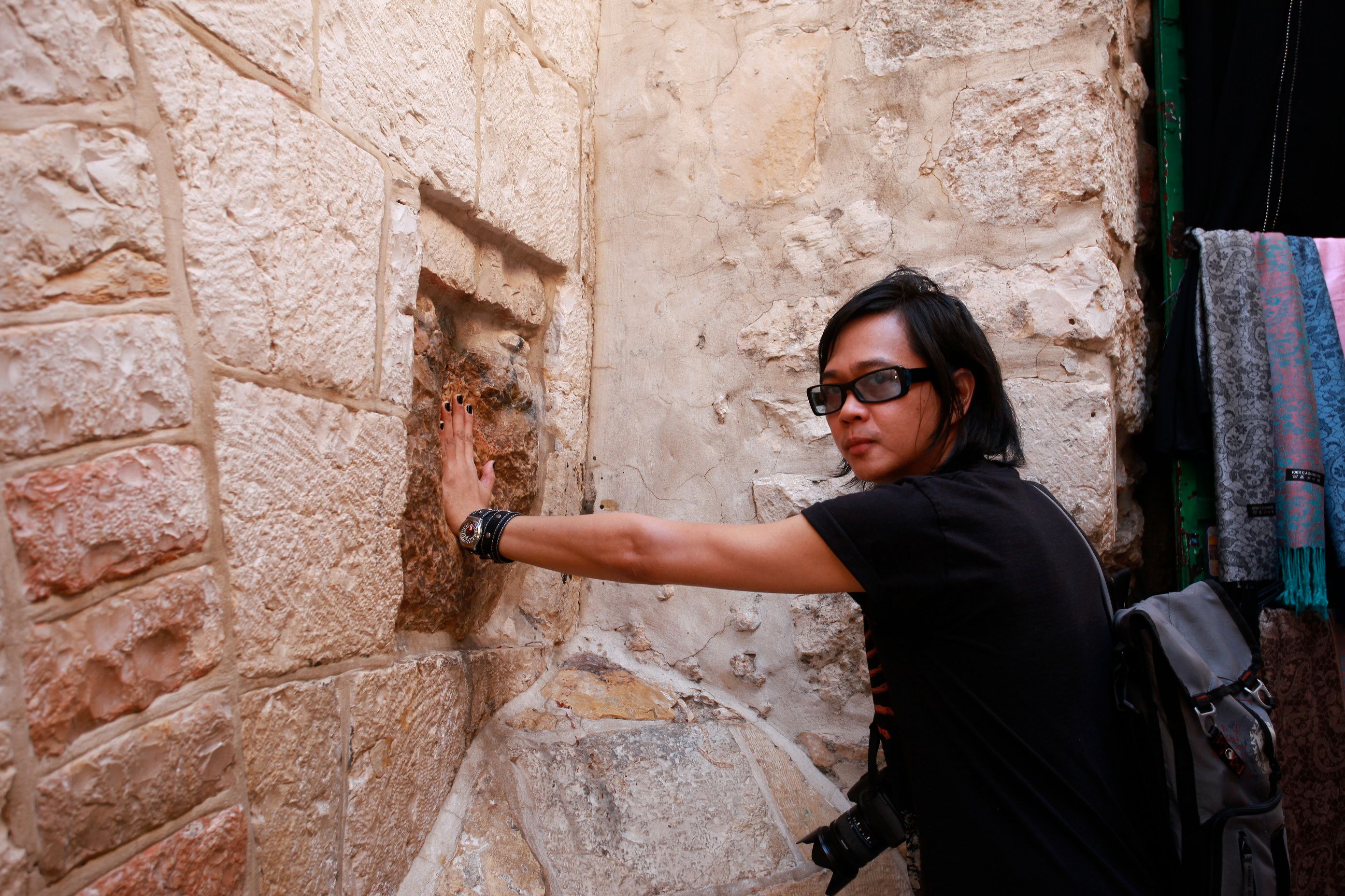 Igan D’Bayan, a camera over his shoulder, presses his hand to a worn stone wall in a narrow Jerusalem alley
