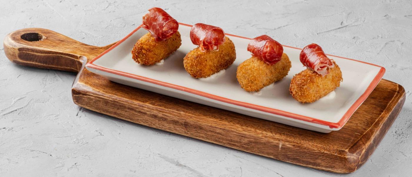 Four golden croquettes draped with curls of Jamon Iberico on a long white plate
