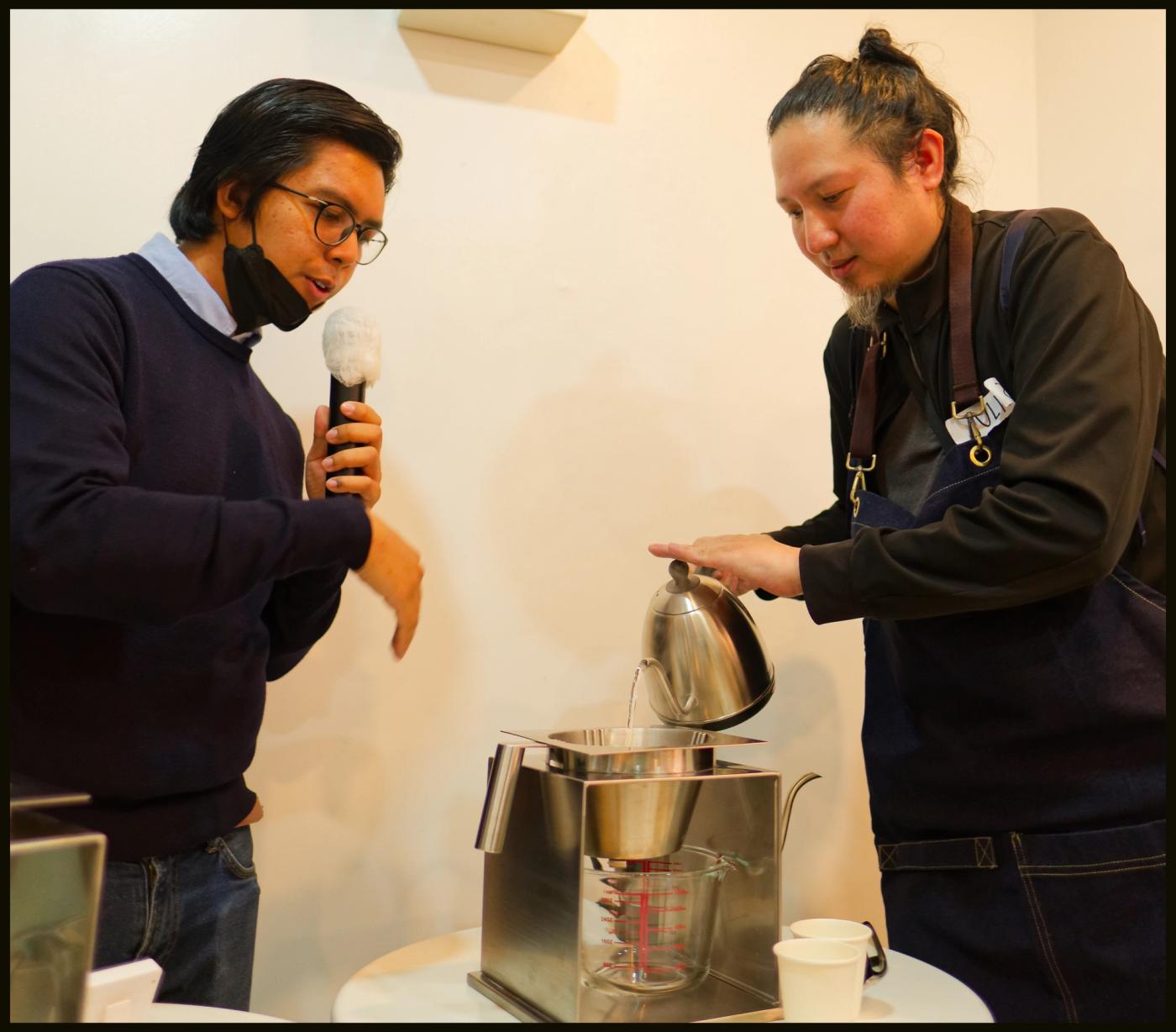 A man in glasses holds a microphone as a barista in a denim apron pours water from a gooseneck kettle into a stainless brewer funnel