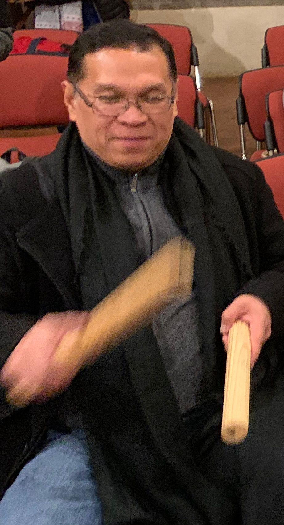 Darwin Gutierrez, in glasses and a dark coat, clacks two wooden percussion sticks while seated among red auditorium chairs