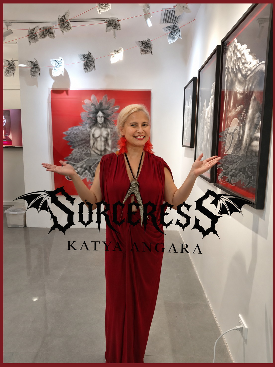 Katya Angara in a red dress and red feather earrings, arms spread before her Sorceress assemblage at The Crucible Gallery, the show’s gothic logotype across the frame