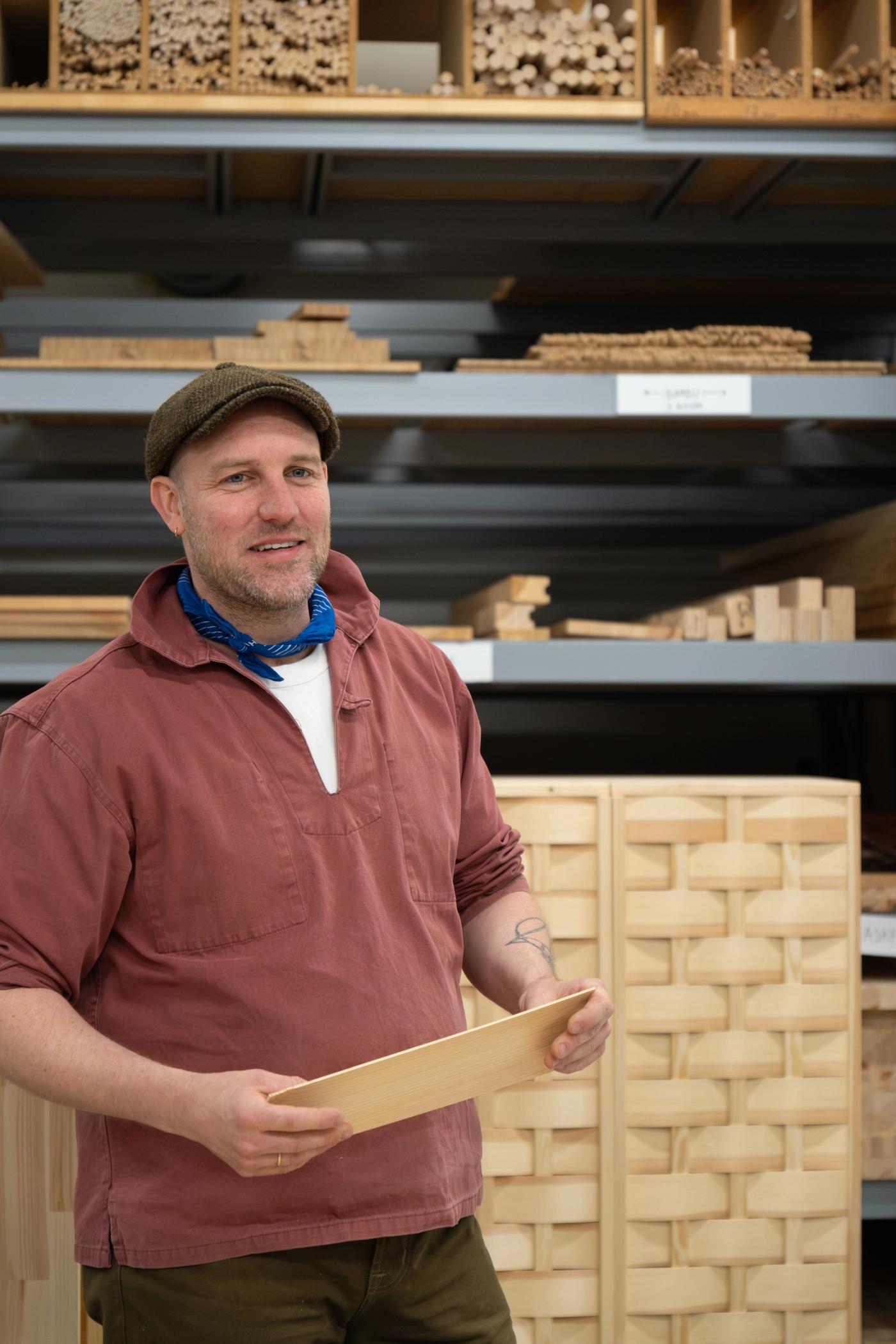 Friso Wiersma in a flat cap holding a wooden shelf panel in front of racks of timber samples